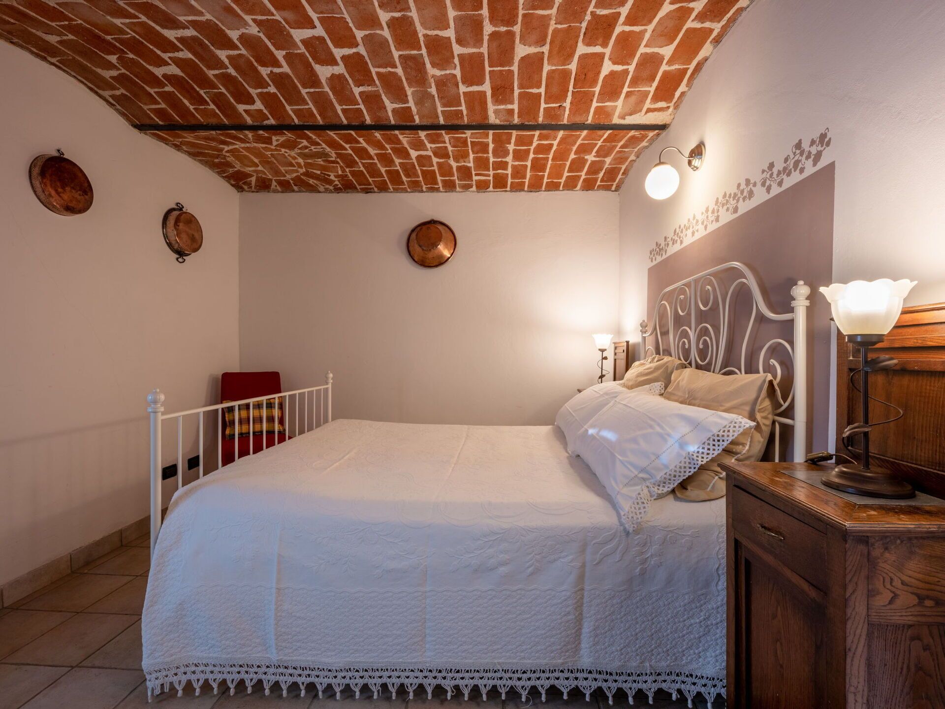 Un dormitorio con techo de ladrillo, cama blanca, mesita de noche con lámpara y cómoda de madera.