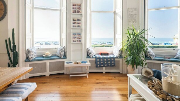 Un coin salon confortable aménagé dans une baie vitrée, avec une table en bois, des bancs rembourrés et une plante en pot.