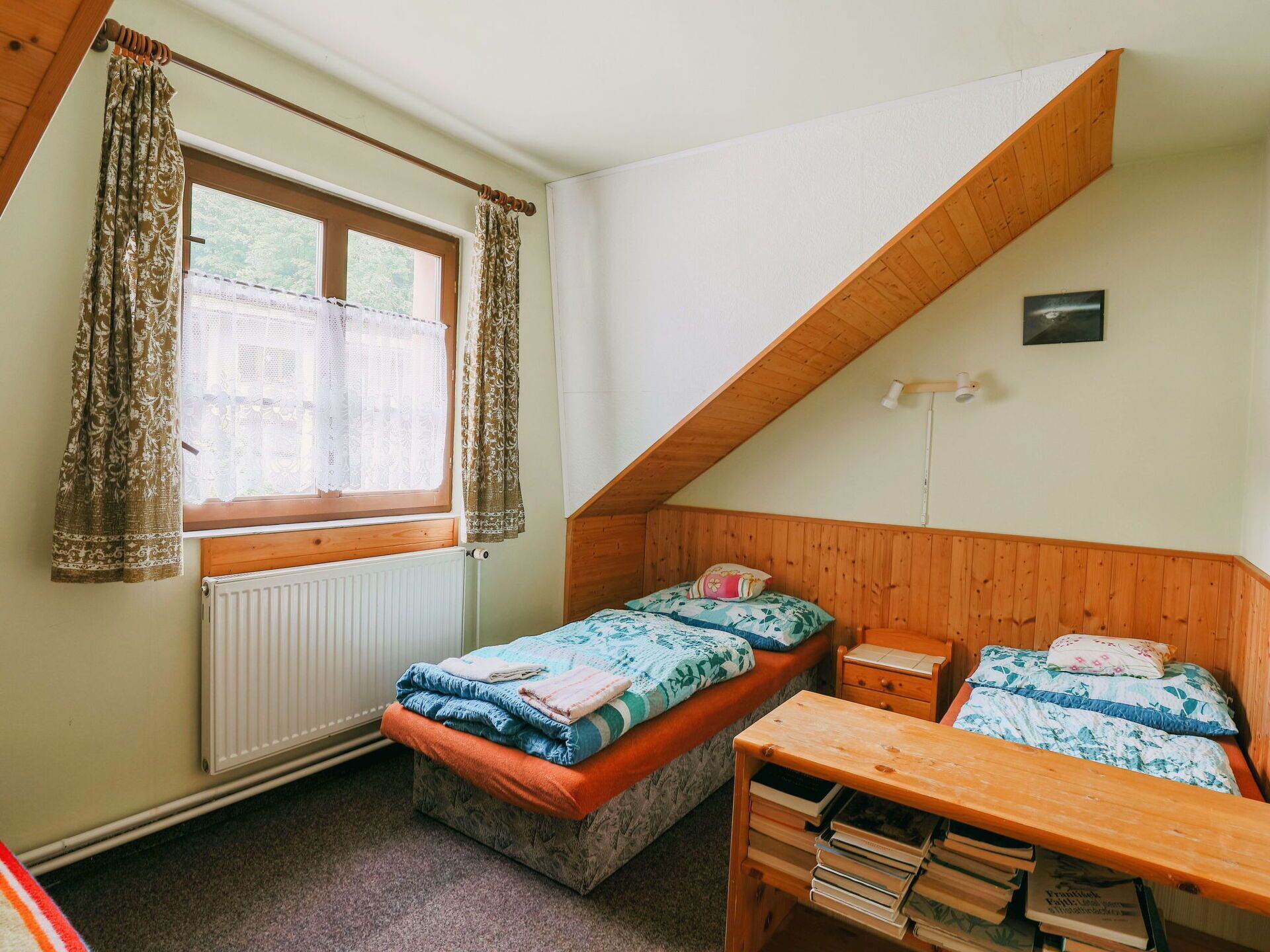 A bedroom with a sloped ceiling, two beds, a wooden desk, and a window with curtains.
