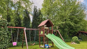 Puinen leikkialue, jossa on liukumäki ja keinut, ja jonka ympärillä on vehreää kasvillisuutta.