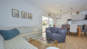 A modern living room with a sofa, armchair, and coffee table. There is a dining area with a table and chairs. The room has a large window and a view of the outdoors.