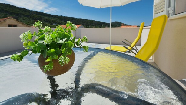 A glass table with a potted plant, a white umbrella, and yellow chairs on a balcony with a view of the hills.