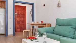 A living room with a green sofa, a white coffee table with a bowl of berries and two wine glasses, a wooden table with a cup and a plate, and a wooden door with a blue frame. - Lovely apartment for 4 people with pool, TV and terrace (Miami Platja)