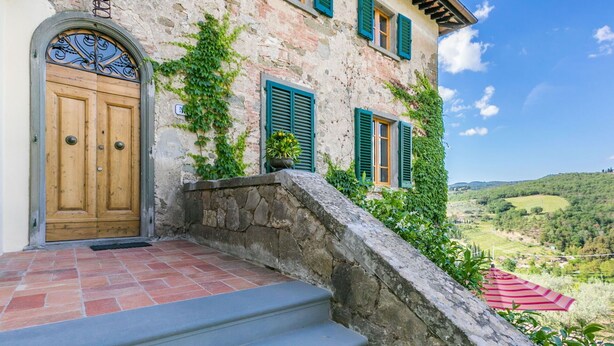 A stone building with a wooden door, green shutters, and a view of a valley.