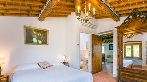 A bedroom with a wooden bed, a chandelier, a painting, and a bathroom visible through an open door.