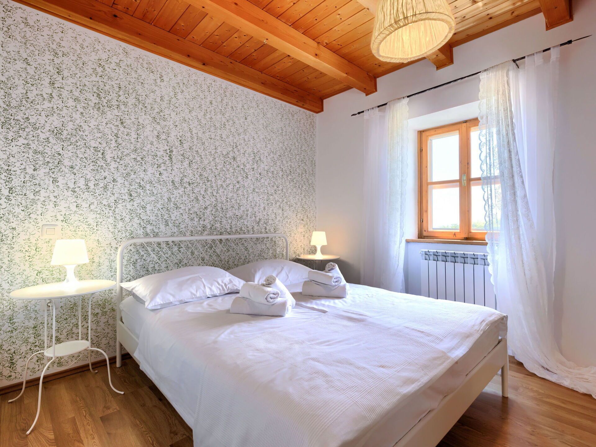 A bedroom with a wooden ceiling, floral wallpaper, a bed with white linens, two bedside tables with lamps, and a window with curtains.