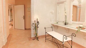 A bathroom with a bench, a mirror, a shelf with toiletries, and a door leading to another room.