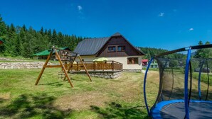 Ein Haus mit einer Holzterrasse, einem Trampolin und einer Schaukel im grünen Garten.