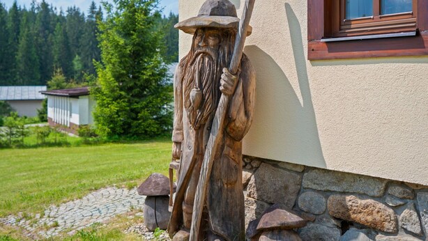 Eine hölzerne Skulptur einer bärtigen Gestalt, die eine Stange hält und an der Außenwand eines Gebäudes befestigt ist.