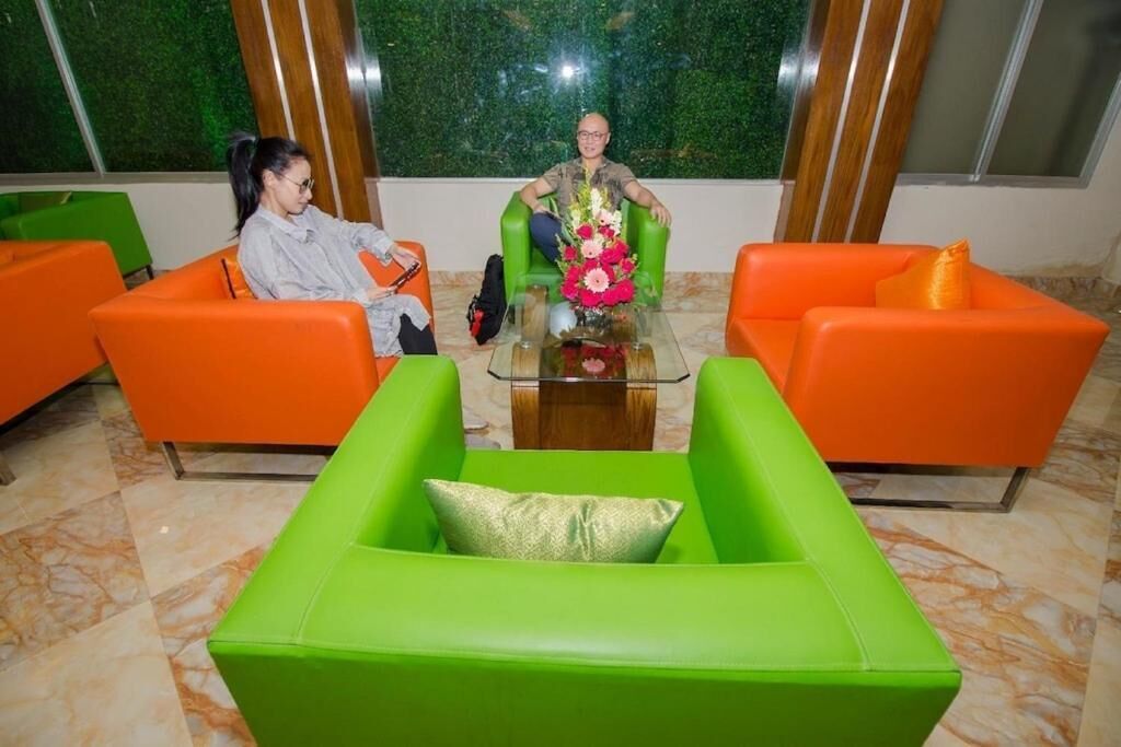 Une pièce comprenant deux fauteuils orange et un fauteuil vert, une table basse en verre agrémentée d’un arrangement floral, et un mur vert orné d’un motif texturé.