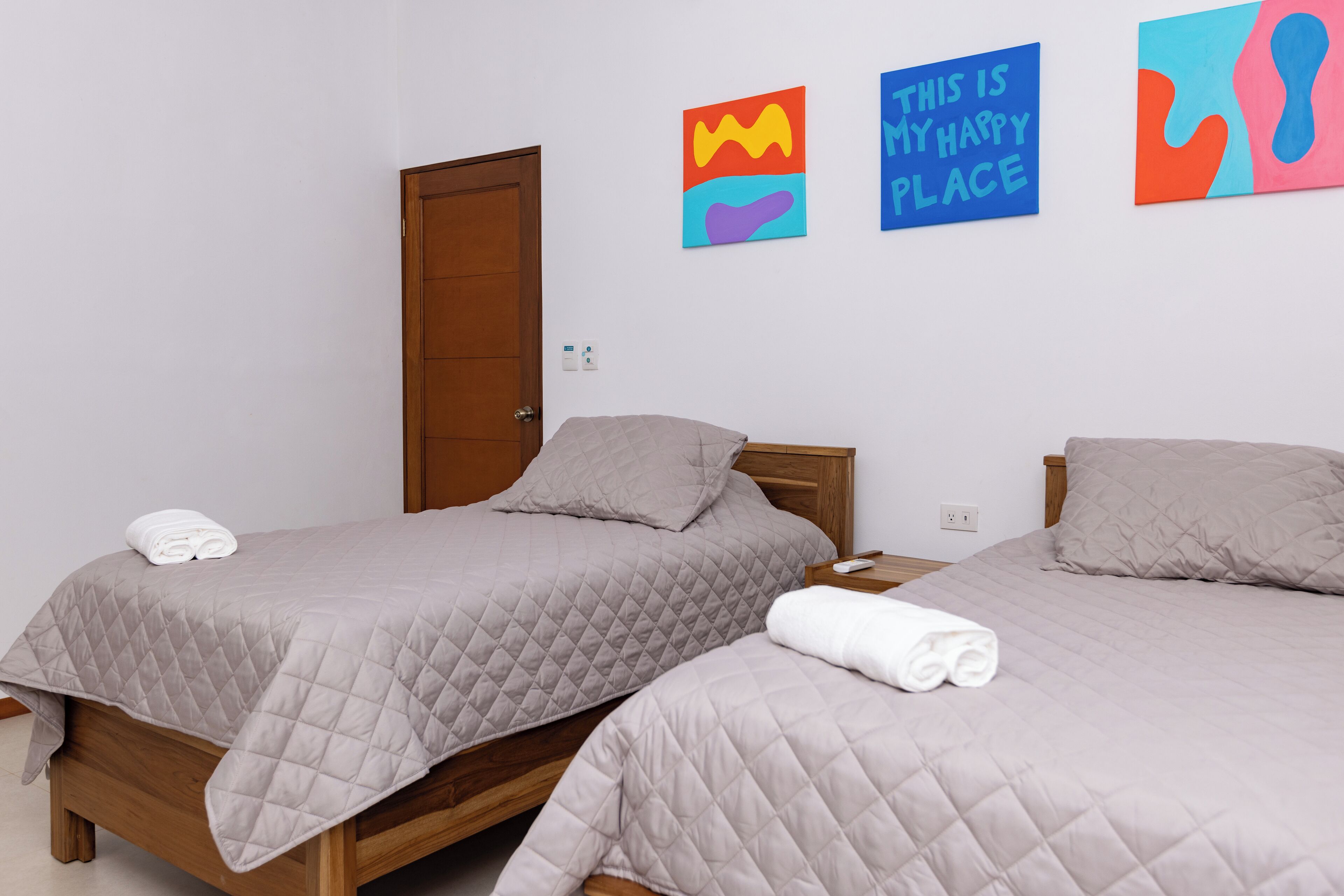 Deux lits avec des couvertures grises et des serviettes blanches, une porte en bois et des œuvres d’art colorées accrochées au mur.