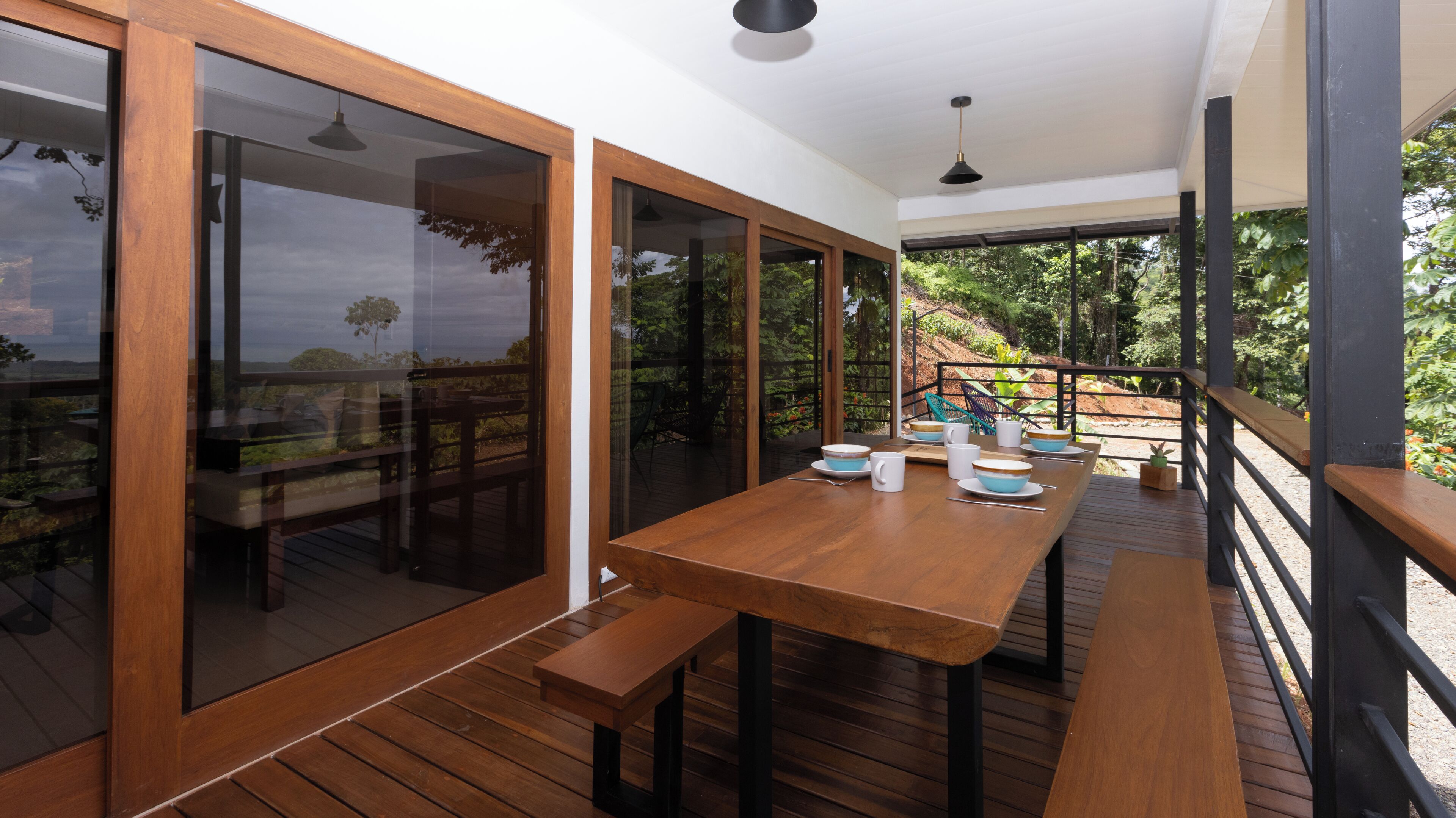 Una terraza de madera con una mesa lista para comer, ventanas de vidrio amplias y vistas a la vegetación.