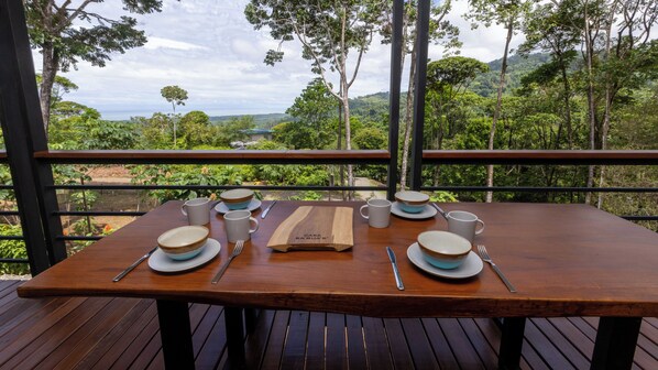 Une table en bois, dressée pour quatre personnes, avec des bols, des assiettes et des couverts, donnant sur une forêt luxuriante et offrant une vue lointaine.