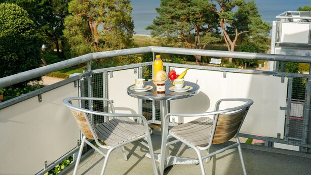 Un balcon avec deux chaises en métal et une petite table ronde, une bouteille de jus d’orange, un saladier de fruits et une tasse. Le balcon donne sur une étendue d’eau et sur des arbres.