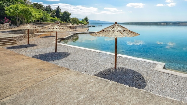 A poolside area with clear blue water, surrounded by pebble ground, and shaded by straw umbrellas. - Beautiful private villa for 4 people with A/C, WIFI, TV and terrace (Zadar)