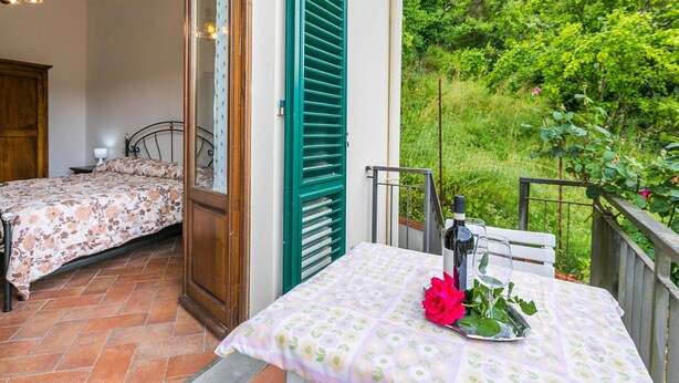 Une chambre à coucher avec un lit, une table de chevet et une fenêtre avec des volets verts. Il y a un balcon avec une table et une bouteille de vin.