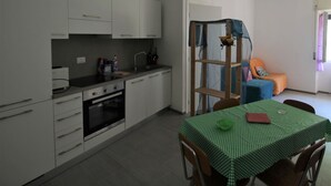 Une cuisine compacte avec des meubles blancs, un four encastré et un coin repas agrémenté d’une nappe à carreaux verts.