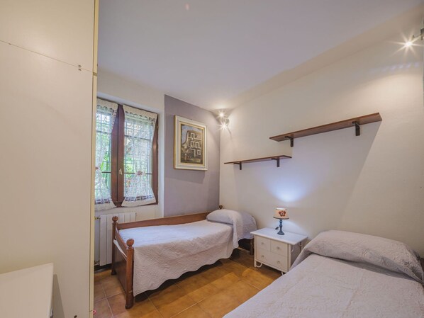 A bedroom with two single beds, a nightstand with a lamp, a window with lace curtains, and a wall-mounted shelf.