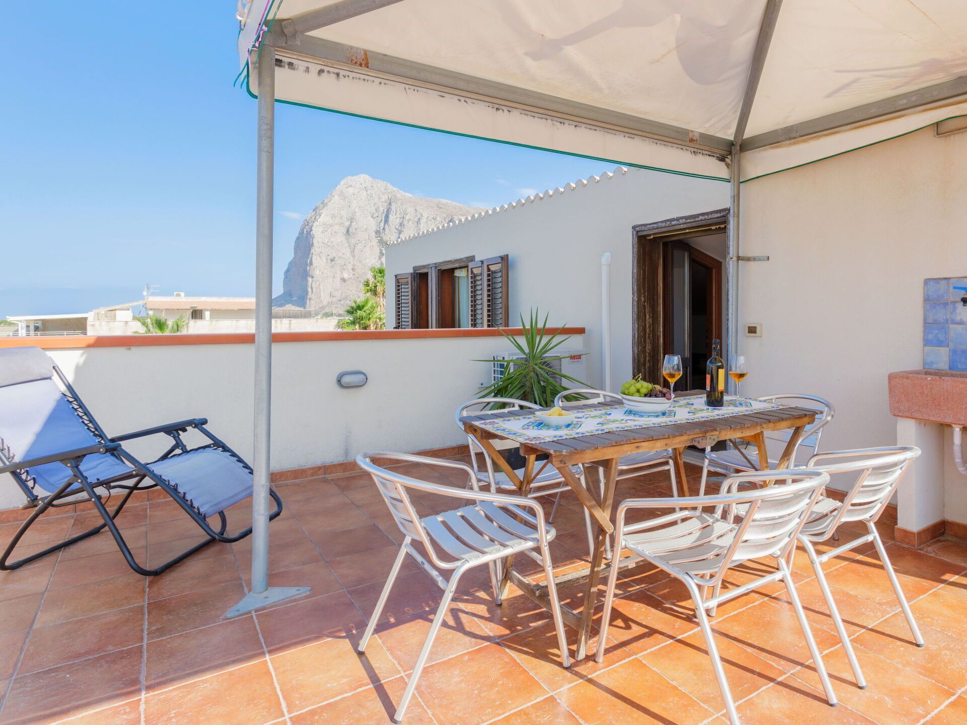 Una terrazza con tavolo apparecchiato per mangiare, sedie, un ombrellone e vista su una montagna.