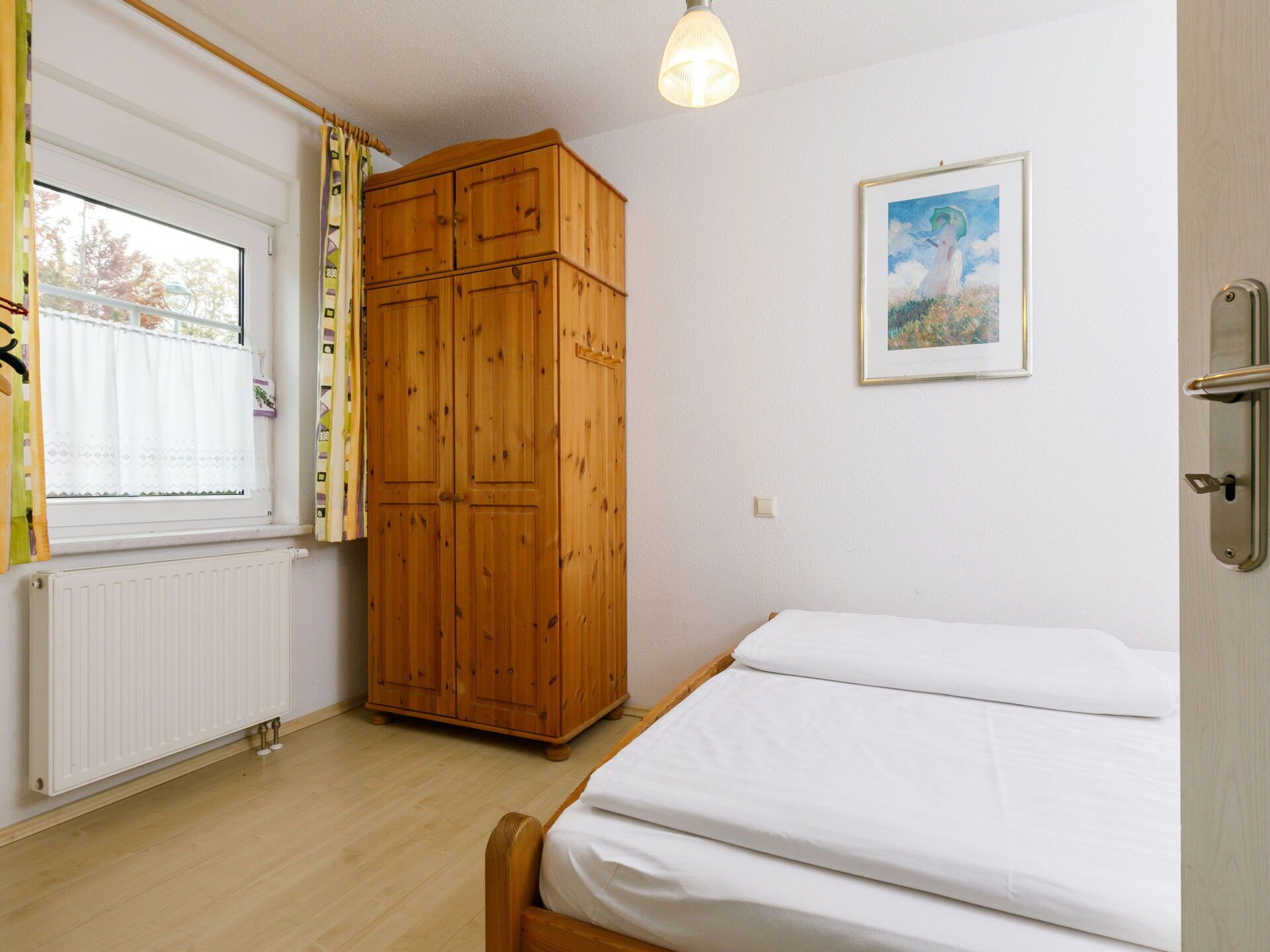 A bedroom with a wooden wardrobe, a bed with white bedding, a radiator, and a window with curtains.