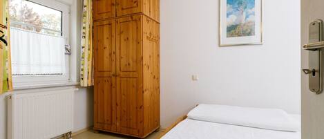 A bedroom with a wooden wardrobe, a bed with white bedding, a radiator, and a window with curtains.