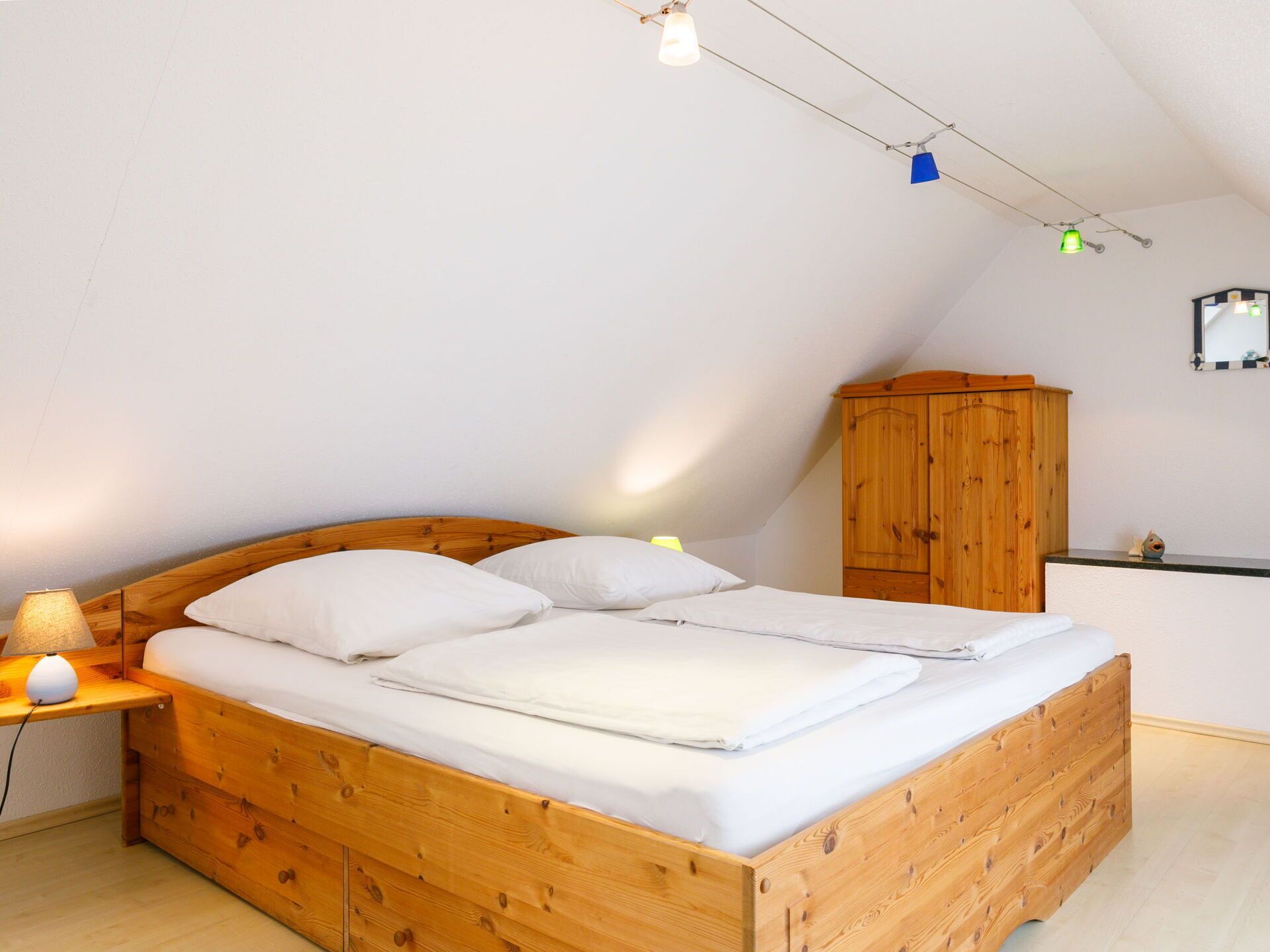 Une chambre individuelle avec un lit en bois, une table de chevet, une lampe, une armoire et un plafond en pente agrémenté de luminaires suspendus.