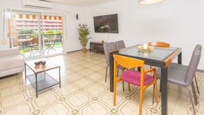 A modern living room with a glass dining table, comfortable seating, a large potted plant, and a view of a residential building through a sliding glass door. - Lovely apartment for 6 guests with pool, WIFI, A/C, TV, balcony and pets allowed (Santa Susanna)