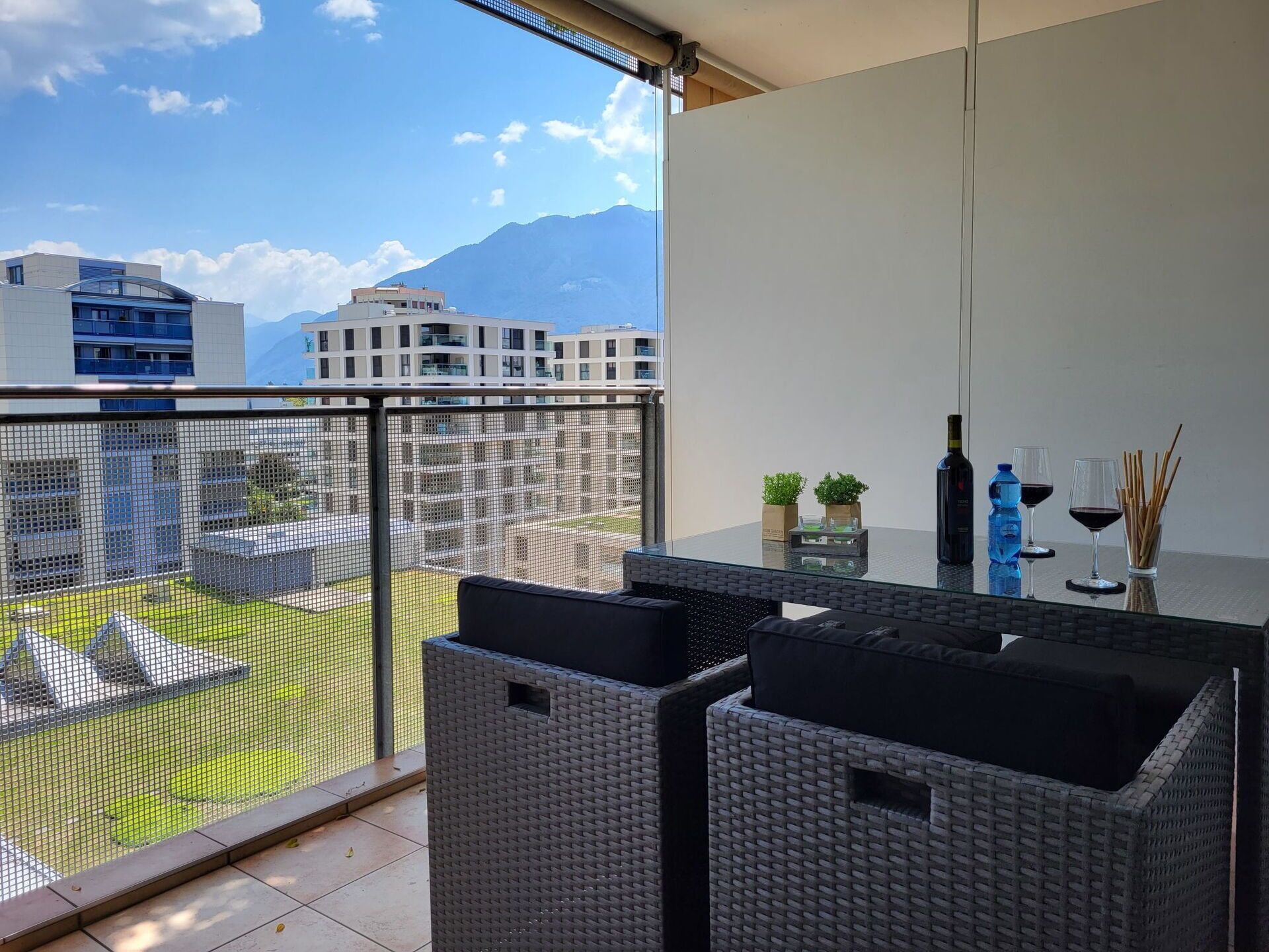 A balcony with wicker furniture, a glass table, and a view of buildings and mountains.