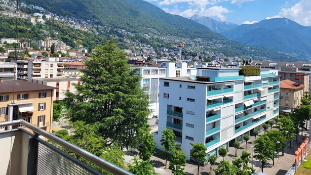 Un edificio moderno con varios pisos y balcones, rodeado de abundante vegetación y montañas al fondo.