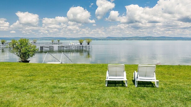 Deux fauteuils de jardin blancs sont placés face à un lac paisible, avec un quai et une zone herbeuse.