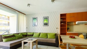 Un salon moderne avec un canapé, une table basse et un coin repas. On y trouve deux tableaux encadrés au mur, une fenêtre avec des rideaux et un coin cuisine doté de meubles en bois et d’une cuisinière.