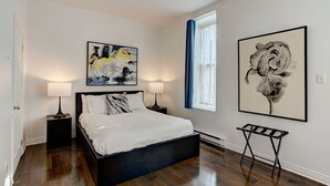 A bedroom with a large bed, two bedside tables with lamps, a window with blue curtains, and two framed artworks on the walls.