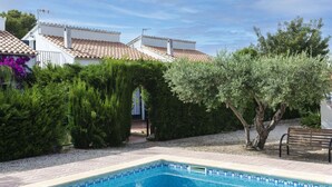 Una piscina con borde de azulejos, rodeada por un jardín con arbustos diversos y una banca.
