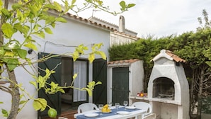 Un patio al aire libre con una mesa preparada para cuatro personas, rodeado de sillas blancas, una pared blanca y un horno de piedra.