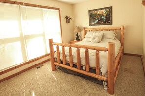 Een slaapkamer met een houten bed, een nachtkastje met een lamp, een raam met jaloezieën en een schilderij aan de muur.