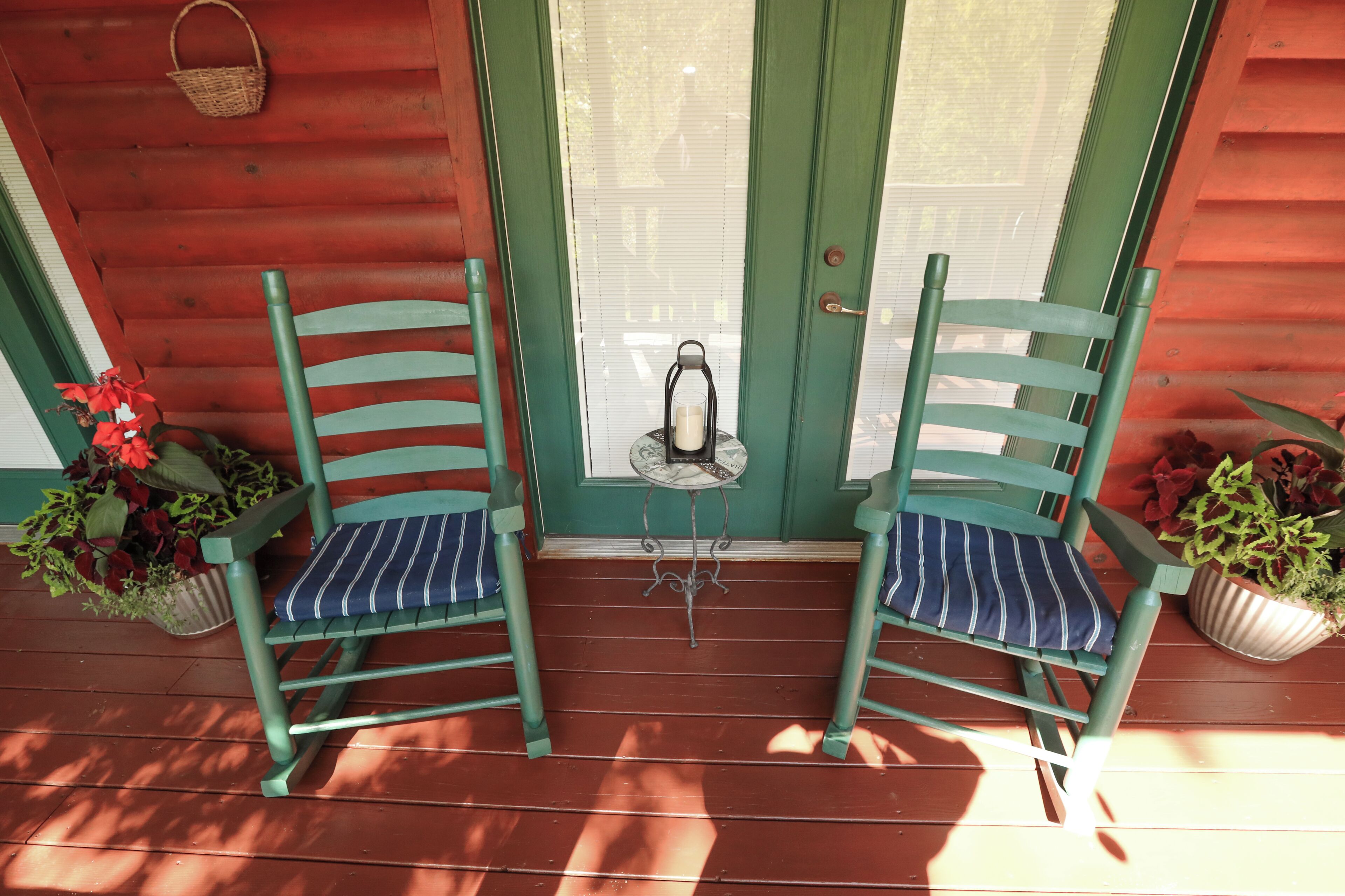 Two green chairs with striped cushions on a wooden porch with a green door, a small table with a lantern, and potted plants.