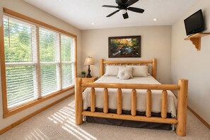 Une chambre avec un lit en bois, un ventilateur de plafond, un téléviseur posé sur une étagère en bois et une grande fenêtre avec des stores.