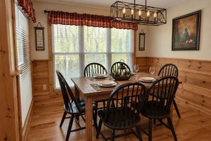 A wooden-paneled dining room with a table set for four, flanked by two windows with blinds, a wall-mounted light fixture, and a framed artwork. - Cardinal's Nest • Sleeps 10 • Near Dollywood, Pigeon Forge & Gatlinburg (Sevierville)