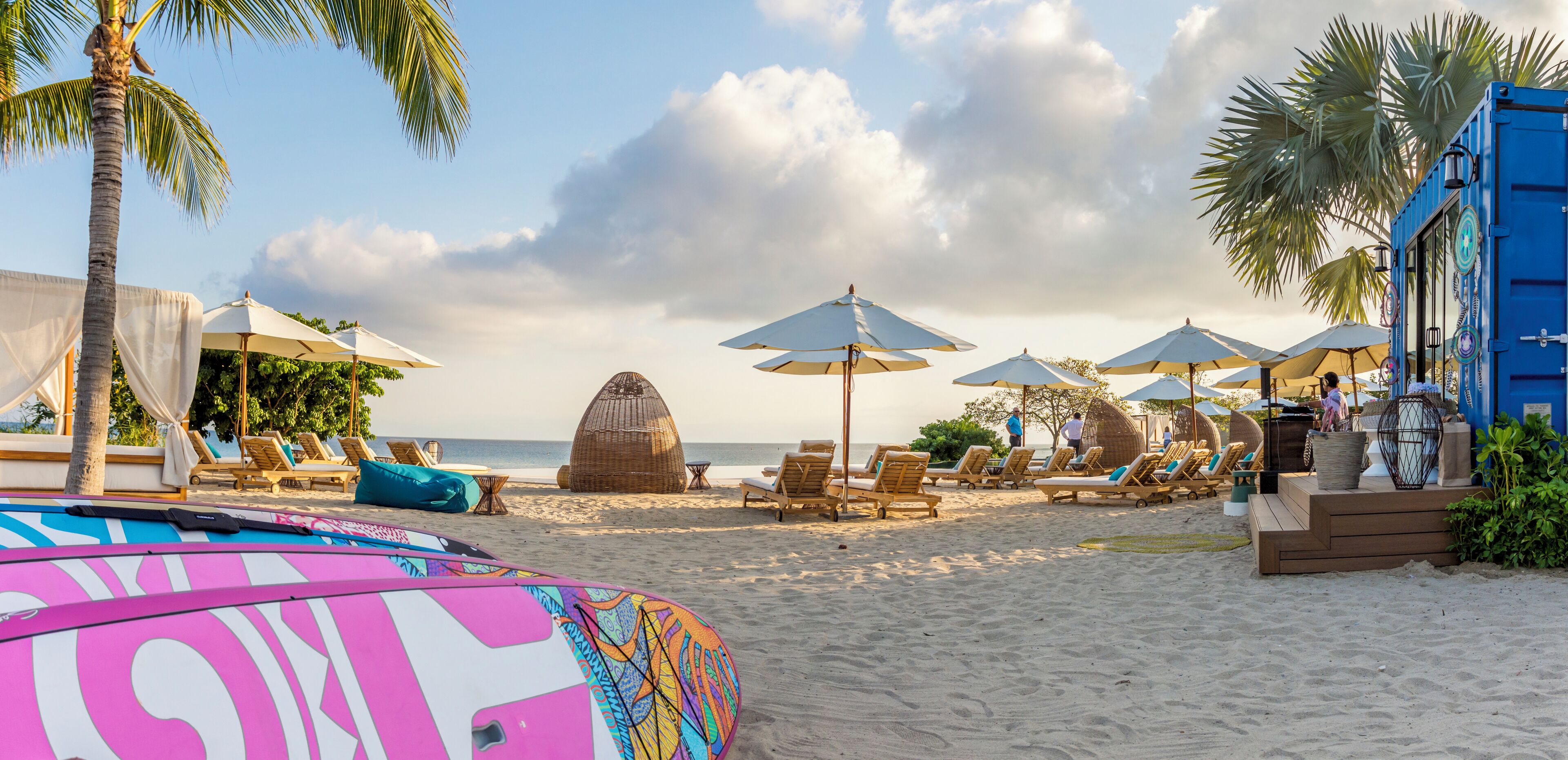 Une plage avec des chaises longues et des parasols, une grande structure tissée et un bâtiment bleu agrémenté d’éléments décoratifs.