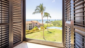 Vista desde una ventana con persianas de madera, que da a una piscina, palmeras y un edificio con techo de tejas.
