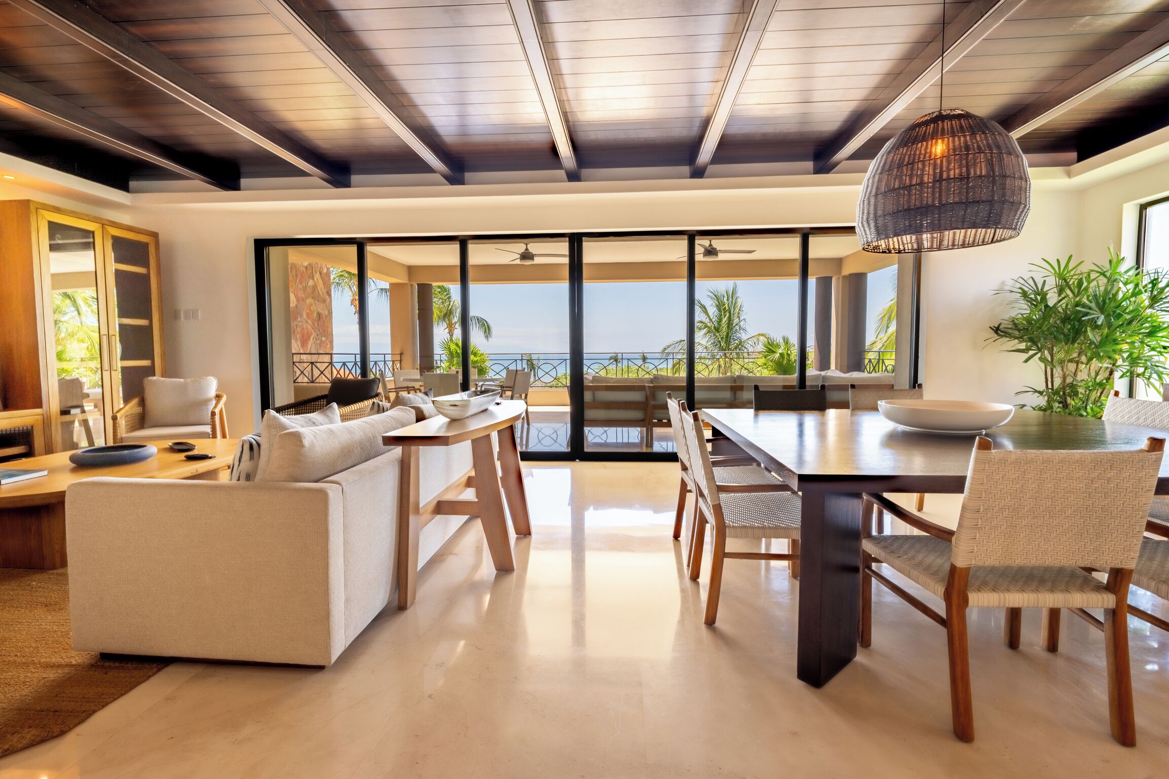A modern living room with a sofa, dining table, and chairs, large windows with a view of the ocean, and a ceiling fan.