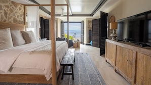 Un dormitorio con una cama grande, un banco de madera, un televisor sobre un mueble de madera y vistas al mar.