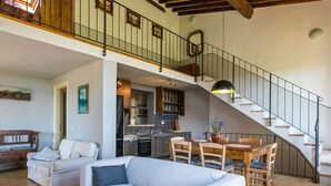A spacious living area with a sofa, a dining table with chairs, a kitchen in the background, and a staircase leading to an upper level.