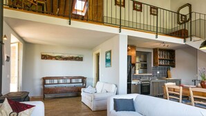 A spacious living area with a high ceiling, a wooden staircase, and a kitchen visible through an open doorway.