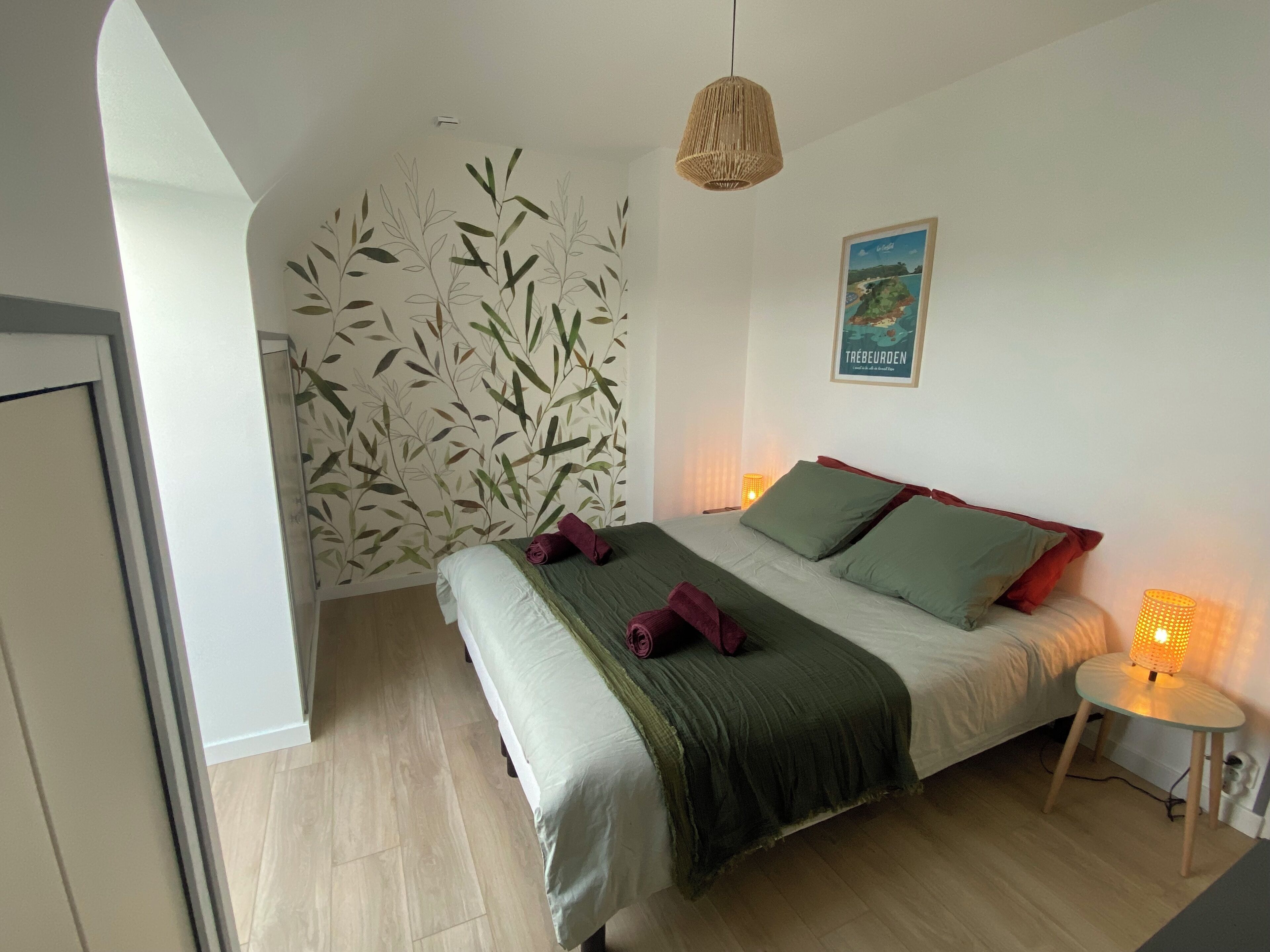 Un dormitorio con cama, pared decorada con motivos de hojas de bambú, lámpara colgante y una mesita de noche con lámpara.