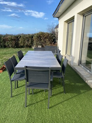 Une table rectangulaire pour l’extérieur, entourée de chaises, placée sur une pelouse bien entretenue, à côté d’un bâtiment doté de grandes fenêtres et avec une haie en arrière-plan.