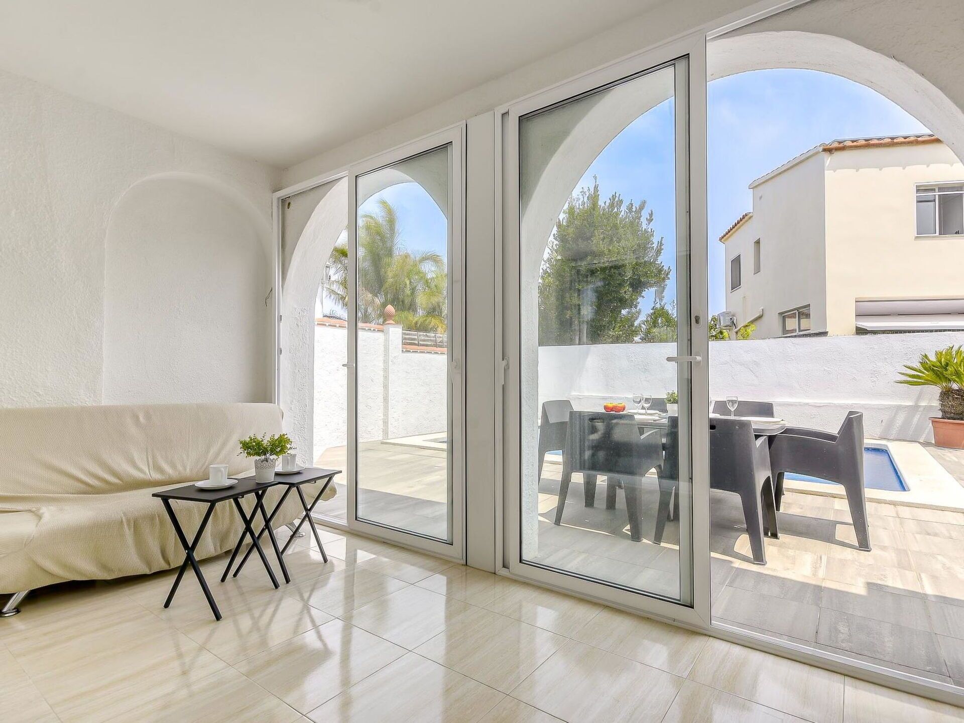 Un moderno salón con un sofá, una mesita y un ventanal de vidrio que da a un patio exterior con piscina y zona de comedor.