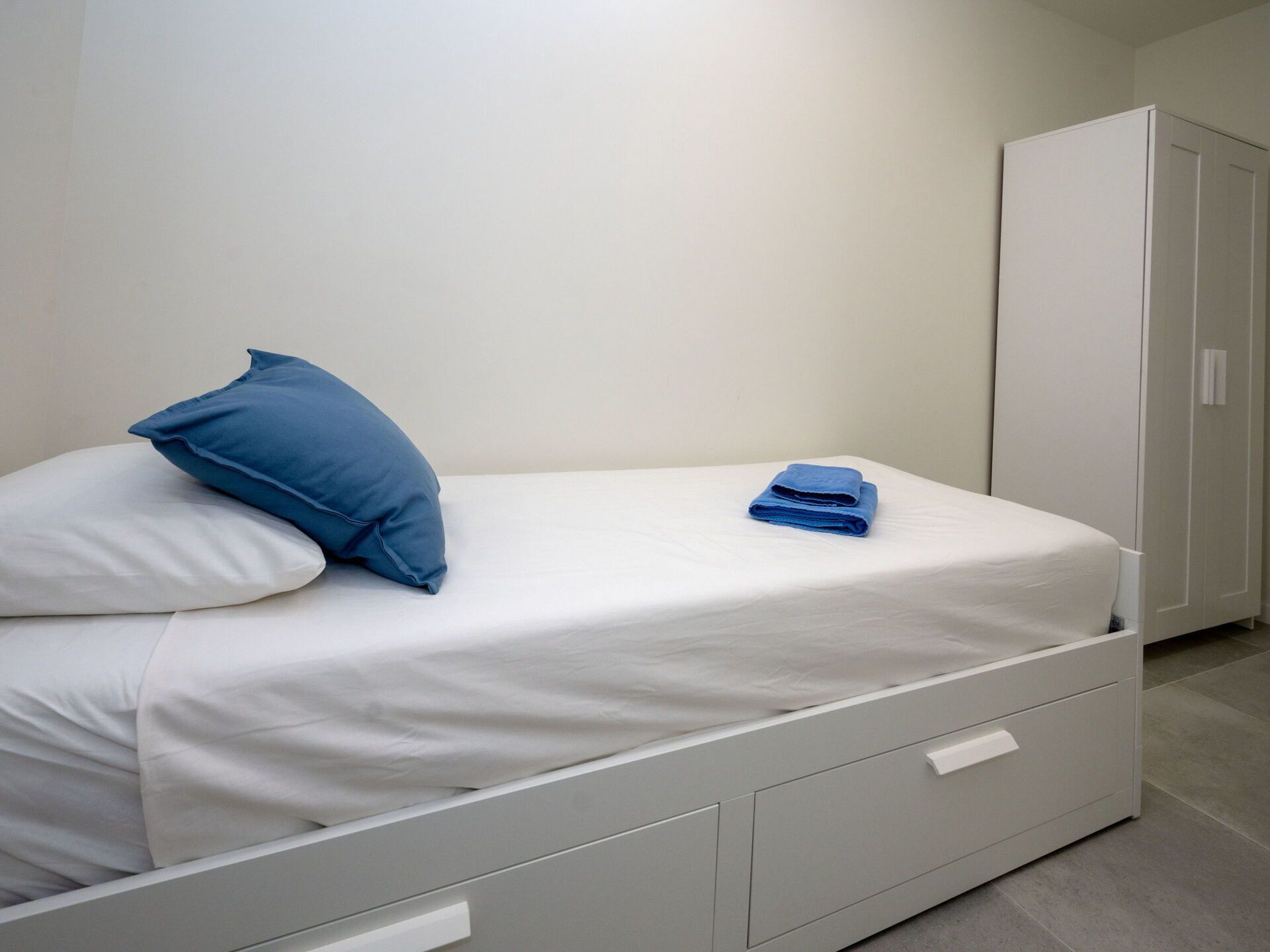 A single bed with a white mattress, a blue pillow, and a folded blue towel. There is a white wardrobe to the right.