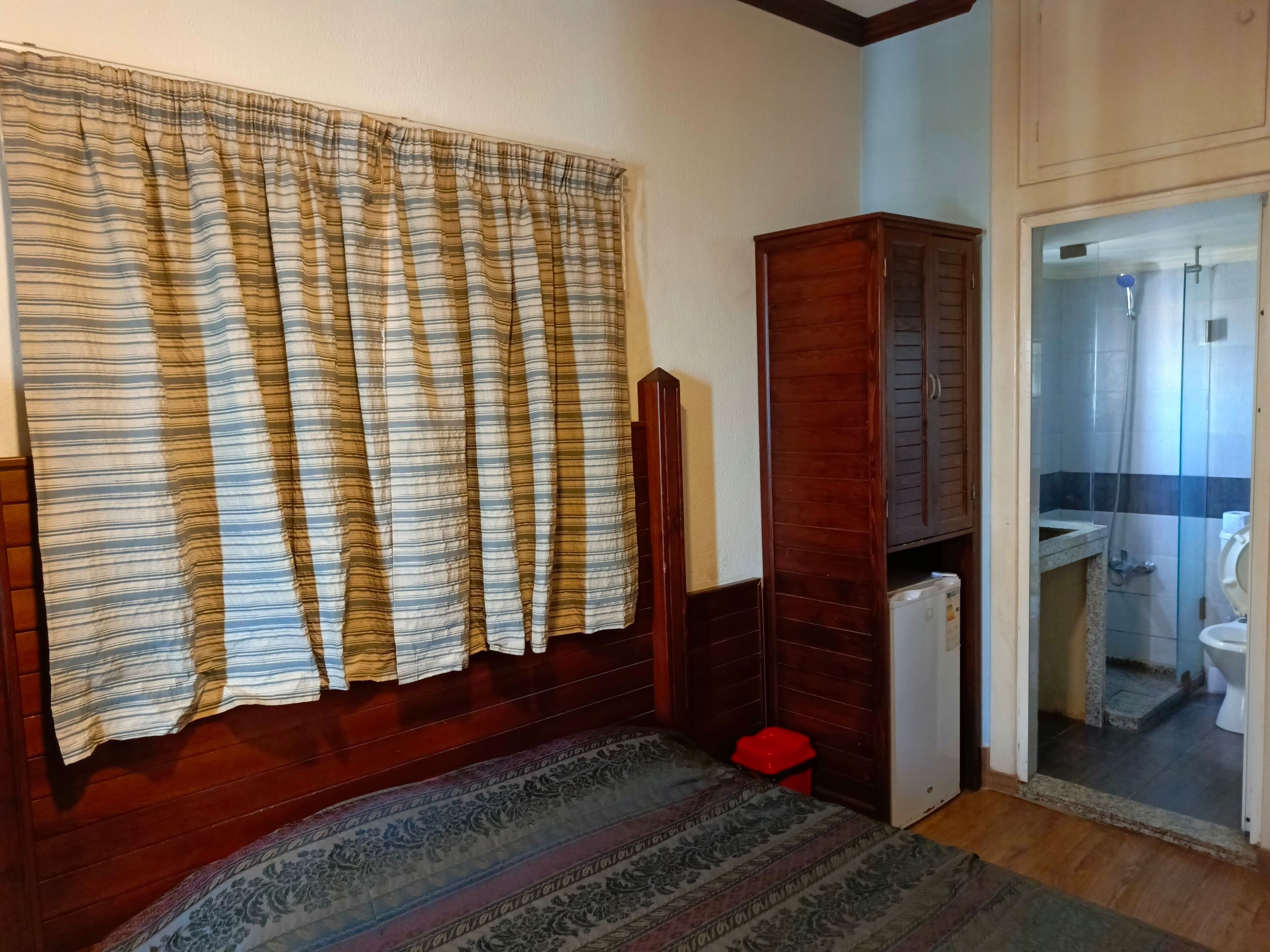 A room with a bed, a wooden wardrobe, a red trash bin, and a bathroom visible through a glass door.