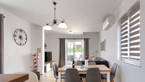 Ett modernt vardagsrum med en matplats, en TV och ett stort fönster med persienner.